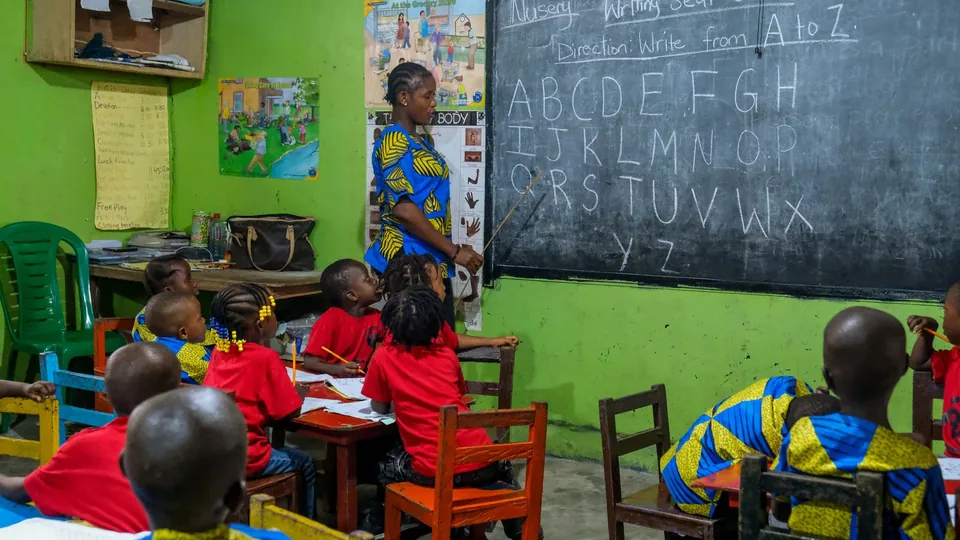 Lehrerin schreibt an die Tafel, Kinder in roter Schuluniform sitzen an Holztischen im Klassenraum.