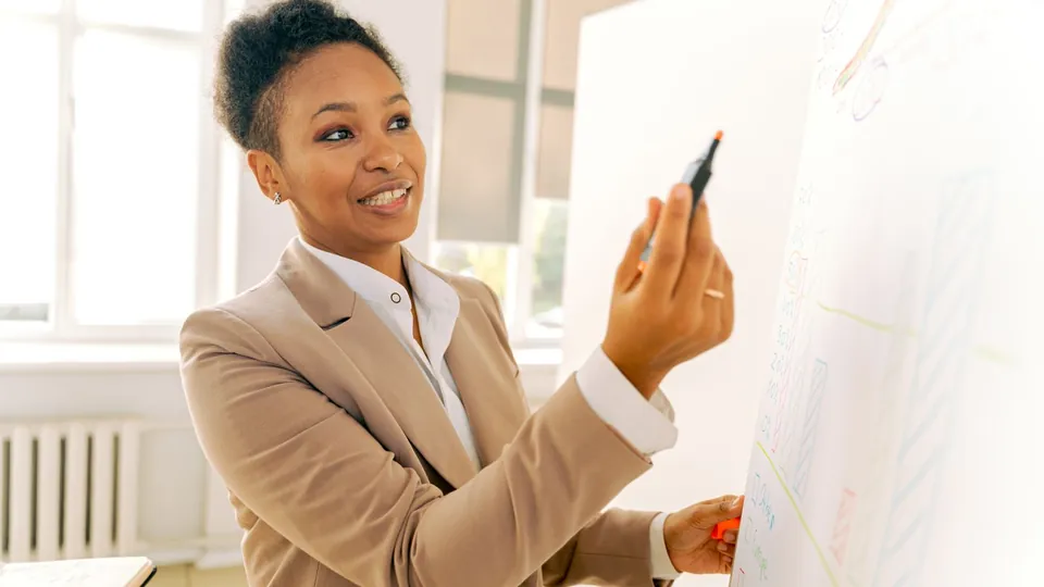 Frau in beigem Blazer schreibt lächelnd mit einem Stift auf ein Whiteboard.